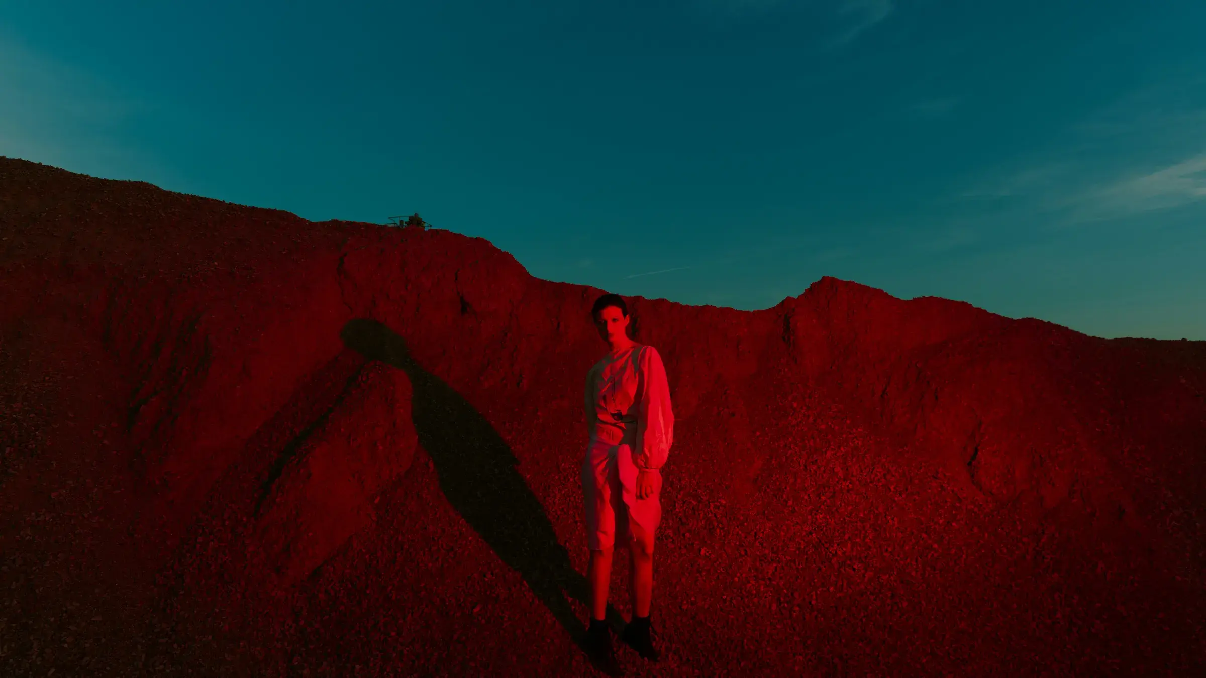 Person standing alone on a red gravel slope under a teal sky